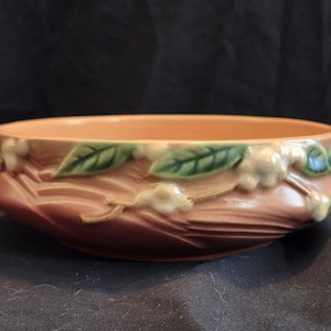 May include: A round, peach-colored ceramic bowl featuring two small handles and a raised, relief-style design of green leaves and white blossoms wrapping around the exterior. The surface has a smooth, matte finish with subtle texture.