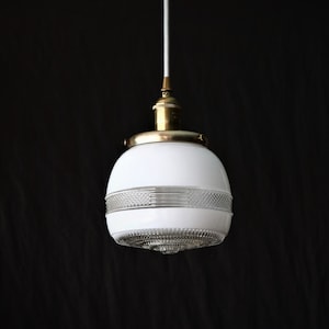 Op de afbeelding: Een witte glazen hanglamp met een messing afwerking en een heldere glazen diffuser. De lamp heeft een geometrisch patroon op het glas.