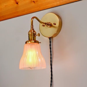 May include: A brass wall sconce with a pink glass shade. The sconce has a simple design and a black cord.