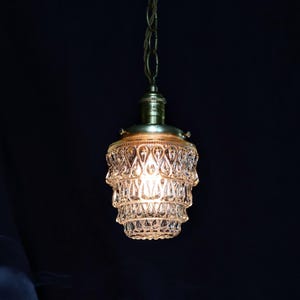 May include: A vintage-style pendant light fitting with a clear glass shade featuring a tiered, faceted design. The shade is illuminated with a warm, white light.