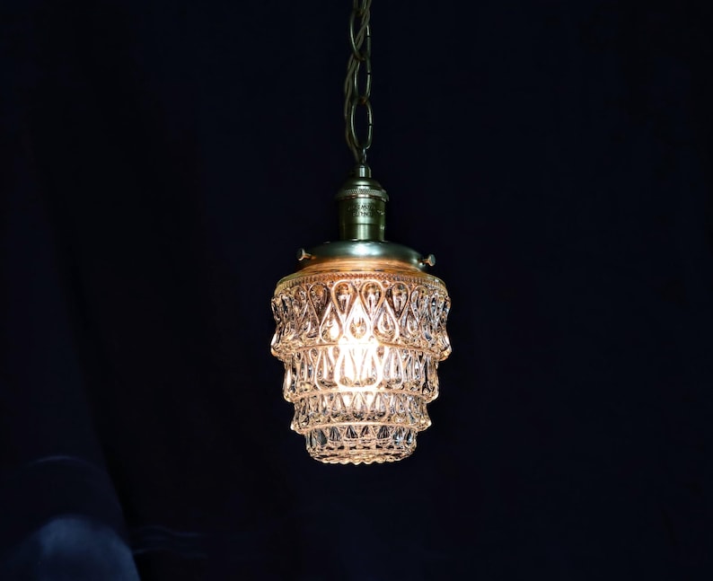 May include: A vintage-style pendant light fitting with a clear glass shade featuring a tiered, faceted design. The shade is illuminated with a warm, white light.