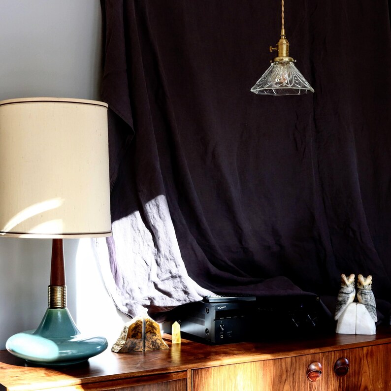 Op de afbeelding: Een tealblauwe tafellamp met een witte kap staat op een houten tafel naast een zwart stereosysteem. Een glazen hanglamp hangt aan het plafond boven de stereo-installatie. Twee decoratieve uil-boekenleggers staan op de tafel naast de stereo-installatie.