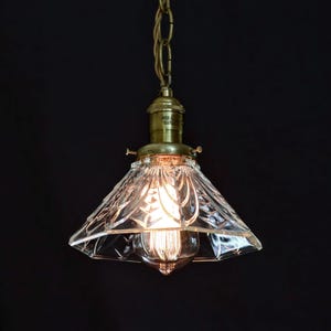 Op de afbeelding: Een messing hanglamp met een heldere glazen kap met een geometrisch patroon. De kap is kegelvormig en heeft een decoratief, facetgeslepen ontwerp.
