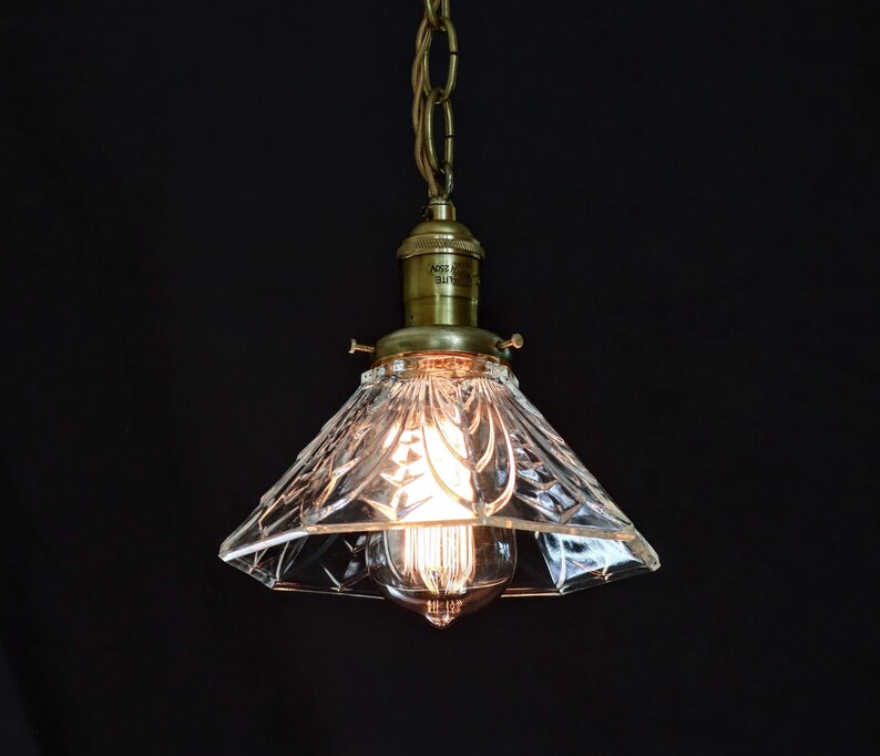 Op de afbeelding: Een messing hanglamp met een heldere glazen kap met een geometrisch patroon. De kap is kegelvormig en heeft een decoratief, facetgeslepen ontwerp.
