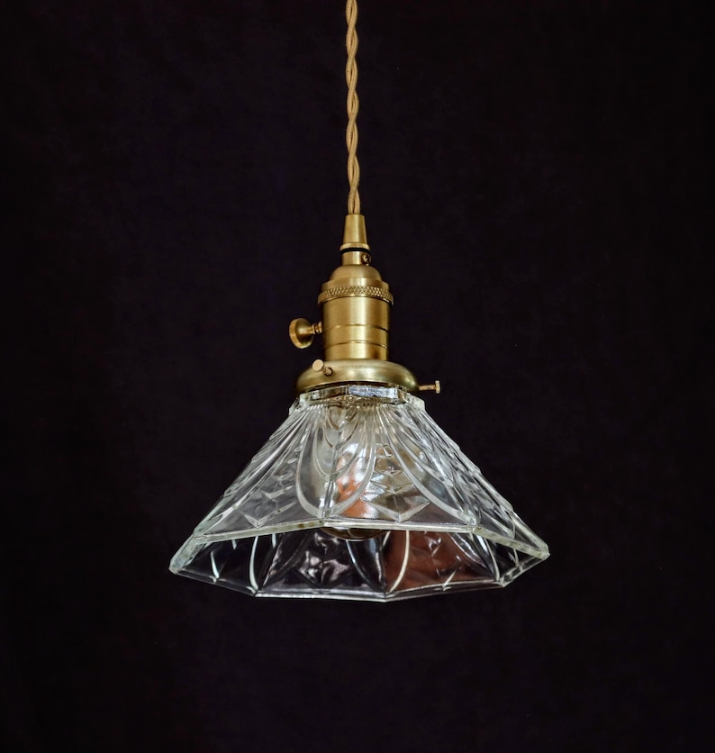 Op de afbeelding: Een messing hanglamp met een heldere, gefacetteerde glazen kap. De kap heeft een geometrisch patroon en hangt aan een gevlochten koord.