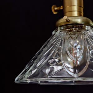 Op de afbeelding: Een transparante glazen lampenkap met een geometrisch patroon. De lampenkap is bevestigd aan een messing lampfitting met een knop.