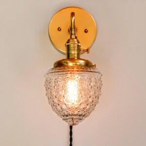 May include: A brass wall sconce with a clear glass shade featuring a textured, bubbled design. The sconce has a simple, elegant design and is perfect for adding a touch of vintage charm to any room.