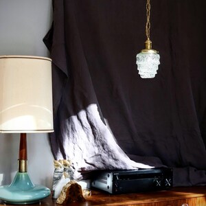 May include: A vintage-style, clear glass pendant light with a brass chain hangs from a dark grey fabric backdrop. The light is illuminating a room with a wooden table and a teal blue lamp with a white shade.