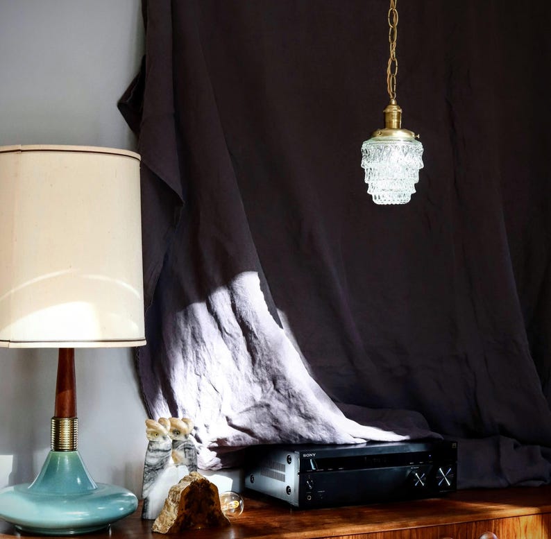 May include: A vintage-style, clear glass pendant light with a brass chain hangs from a dark grey fabric backdrop. The light is illuminating a room with a wooden table and a teal blue lamp with a white shade.