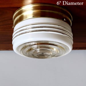 Op de afbeelding: Een plafondlamp met een diameter van 15 cm, een messing rand en een witte glazen kap. De kap heeft een gelaagd, getextureerd ontwerp.