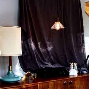 Op de afbeelding: Een vintage-stijl tafellamp met een witte kap en een tealblauwe basis. De lamp staat op een houten tafel met een zwarte stereo-ontvanger en twee uil-boekenleggers.