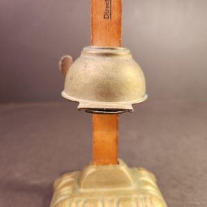 May include: A vintage brass-colored object with a bell-shaped top and a wooden stick. The base has an ornate design. The word "Direct" is printed on the wooden stick. The object is likely a tool or decorative item.