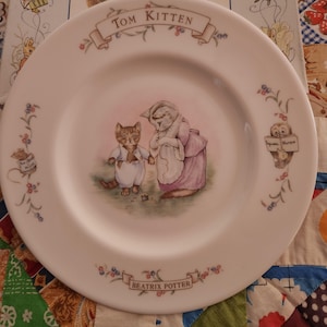 May include: White ceramic plate featuring a Tom Kitten illustration by Beatrix Potter. The plate has a central image of two cats, one in a pink dress and the other in a white outfit. Floral and text details are around the rim.