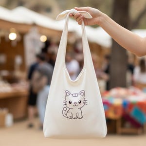 May include: Cream-colored tote bag with a cartoon cat illustration. The bag has long handles tied at the top, held by a hand. The cat illustration is a simple line drawing with a smiling face and whiskers.