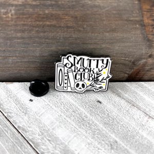 May include: A black and white enamel pin with the text "Smutty Book Club". The pin features a skull, books, and botanical elements. A black pin back is next to the pin on a wooden surface.