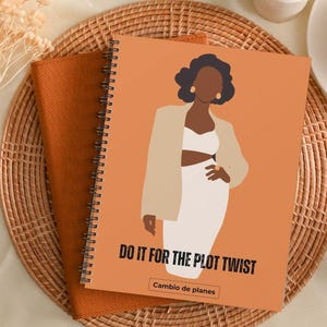 Puede incluir: Cuaderno de espiral con una cubierta naranja que presenta una ilustración de una mujer con blazer y vestido. El texto "DO IT FOR THE PLOT TWIST" está impreso en la cubierta. El cuaderno está sobre un mantel individual tejido con té y lápices.