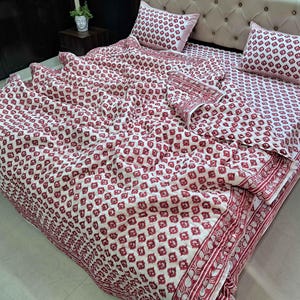 May include: A white and red floral patterned bedding set. The set includes a comforter, two pillowcases, and a bedspread. The comforter and pillowcases feature a repeating pattern of small red floral shapes. The bedspread has a decorative border with a floral design.
