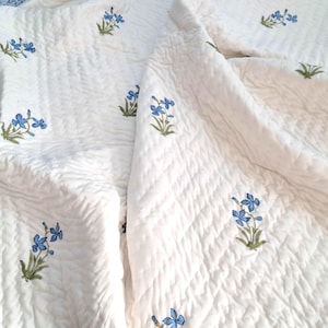 May include: White quilted bedspread with a floral pattern. The bedspread features a repeating design of blue flowers with green stems and leaves. The fabric has a textured, rippled appearance.