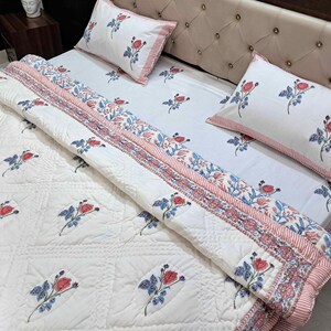 Peut inclure: Ensemble de literie floral blanc comprenant une couette, deux taies d'oreiller et un drap-housse. L'ensemble présente un motif de roses rouges et de feuilles bleues. La couette et les taies d'oreiller ont une bordure rayée rose.