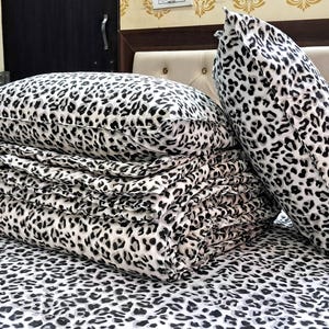 May include: A bed set featuring a black and white leopard print pattern. The set includes a folded blanket, two pillows, and a fitted sheet. The pillows are a standard rectangular shape and a long bolster pillow. The bedding is neatly arranged on a bed.