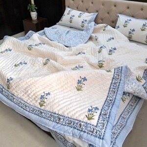 May include: A cream-colored quilt and pillow set with a blue floral pattern. The quilt has a quilted texture and a blue border with a floral design. Two matching pillowcases are visible on the bed.