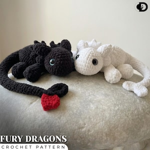 May include: Two crocheted dragon toys, one black and one white, are displayed. The black dragon has a red tail accent. The toys have large, round eyes. The text "FURY DRAGONS CROCHET PATTERN" is at the bottom of the image.