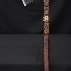 May include: Axe with a dark blade and a wooden handle. The axe head features decorative carvings. The handle is wrapped in leather and has additional carved details and metal accents. The axe is displayed against a black background.