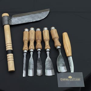 May include: A collection of woodworking tools, including a large drawknife and several chisels, arranged on a black surface. The tools have wooden handles and metal blades. A business card with the text "EMPIRE CUTLERY" is also present.