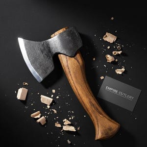May include: A small axe with a wooden handle and a dark metal head. The axe is surrounded by wood shavings and a business card that reads "EMPIRE CUTLERY". The axe is likely used for woodworking or outdoor activities.