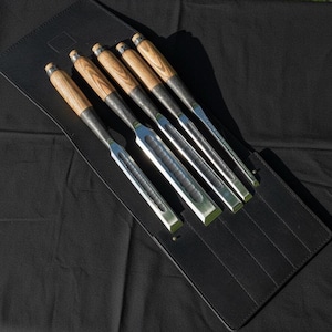 May include: A set of six wood chisels with wooden handles and metal blades, presented in a black leather tool roll. The chisels feature natural wood grain handles and polished metal blades, ideal for woodworking.