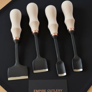 May include: Four chisels with light wood handles, copper accents, and dark gray blades are displayed on a black surface. The chisels have different blade shapes. A business card with "EMPIRE CUTLERY" is also present.