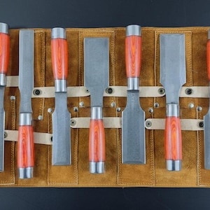 Könnte beinhalten: Ein Satz Holzmeißel mit orangefarbenen und silberfarbenen Griffen, präsentiert in einer braunen Leder-Werkzeugrolle. Die Meißel haben silberne Klingen und werden von Lederriemen gehalten. Die Werkzeugrolle wird mit zwei Schnallen verschlossen.