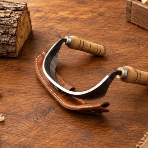 May include: A drawknife featuring a curved metal blade and two cylindrical wooden handles with black ring details. The tool is partially encased in a brown leather sheath with white stitching, designed for woodworking and shaping timber.