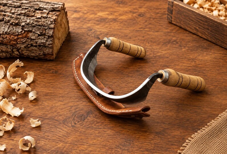 May include: A drawknife featuring a curved metal blade and two cylindrical wooden handles with black ring details. The tool is partially encased in a brown leather sheath with white stitching, designed for woodworking and shaping timber.