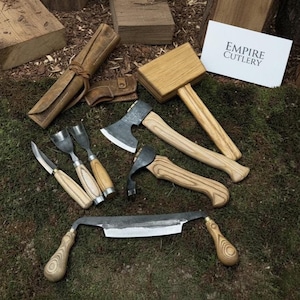 Puede incluir: Colección de herramientas para trabajar la madera, que incluye un hacha, un mazo, un cuchillo de tracción y cuchillos para tallar, con mangos de madera y hojas de metal. También hay un rollo de herramientas de cuero y una tarjeta con "EMPIRE CUTLERY".