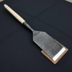May include: A handcrafted metal spatula with a light wood handle and a dark metal blade. The spatula rests on a small, square leather pad with white stitching. The handle is approximately 12 inches long.