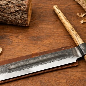 May include: A hand tool featuring a long, rectangular metal blade with a dark, textured finish and a sharp edge, attached to a cylindrical wooden handle with grooved detailing. The blade rests on a rectangular brown leather sheath with stitching.