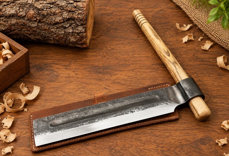 May include: A hand tool featuring a long, rectangular metal blade with a dark, textured finish and a sharp edge, attached to a cylindrical wooden handle with grooved detailing. The blade rests on a rectangular brown leather sheath with stitching.