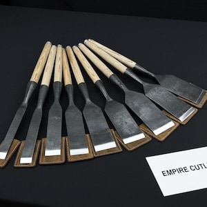 Puede incluir: Un conjunto de formones con mangos de madera y hojas de metal oscuro, dispuestos en abanico. Las hojas tienen forma rectangular y están colocadas sobre almohadillas de cuero y blancas. Las herramientas se exhiben sobre una superficie negra. Se ve una tarjeta blanca con "EMPIRE CUTLERY".