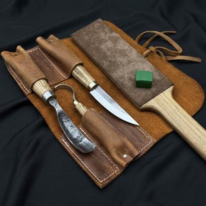 May include: A wood carving set with three tools, including a knife and a spoon carving tool, nestled in a brown leather sheath. A sharpening strop and a green honing compound are also included. The tools have wooden handles and metal blades.