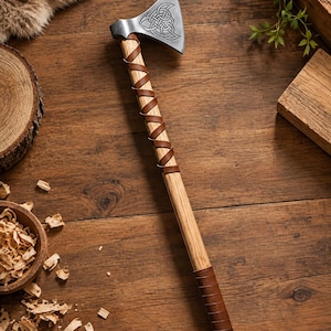May include: A hand axe with a silver-colored metal head featuring an engraved Celtic knot design. The handle is made of light-colored wood, wrapped with a brown leather cord in a spiral pattern, and finished with a brown leather grip at the base.