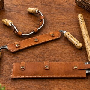 May include: Two drawknives with wooden handles and metal blades, each featuring a tan leather sheath with metal snaps. One drawknife is curved, while the other is straight. The tools are designed for woodworking and are shown with wood shavings.