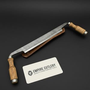 May include: A hand-forged draw knife with a wooden handle and a leather sheath. The blade is made of steel and has a curved shape. The knife is designed for woodworking and is a quality hand-forged tool.