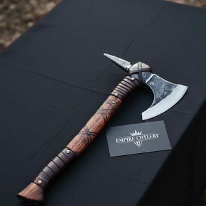 Puede incluir: Un hacha artesanal con mango de madera y hoja de metal. El mango está envuelto en cuero y adornado con tachuelas metálicas decorativas. Una tarjeta de visita con el texto "EMPIRE CUTLERY" está junto al hacha.