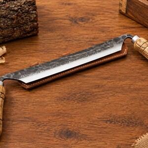 May include: A drawknife with a long, straight, dark hammered metal blade and two light brown wooden handles with black ring details. The blade is partially inserted into a brown leather sheath, featuring a sharp, polished cutting edge.