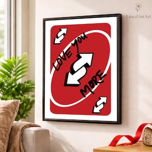 May include: Framed art print featuring a red card design with the words "Love You More" in black script. The card has a white oval in the center with a white arrow. The print is in a black frame and hangs on a white wall.