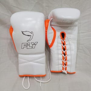 Può includere: Un paio di guantoni da boxe bianchi con accenti arancioni brillanti. Un guantone presenta un logo nero di una figura alata sopra la scritta FLY. L'altro guantone mostra un sistema di chiusura a lacci bianchi con bordi arancioni.