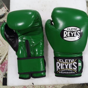 Könnte beinhalten: Paar grüne Boxhandschuhe mit schwarzen Akzenten. Die Handschuhe tragen das Logo "CLETO REYES" in Weiß auf schwarzem Grund. Sie bestehen aus glänzendem Material und sind für den professionellen Einsatz konzipiert.