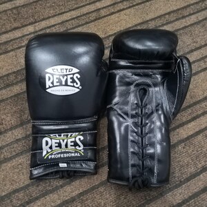 Peut inclure: Une paire de gants de boxe en cuir noir avec des coutures blanches. Les gants portent le logo "CLETO REYES" en blanc et le texte "HECHO EN MEXICO". La zone du poignet porte le texte "REYES PROFESIONAL".