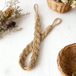 May include: Braided jute rope door pull with loop handles. The natural brown rope is arranged in a decorative pattern. Dried flowers and woven baskets are in the background, suggesting a rustic or farmhouse style.